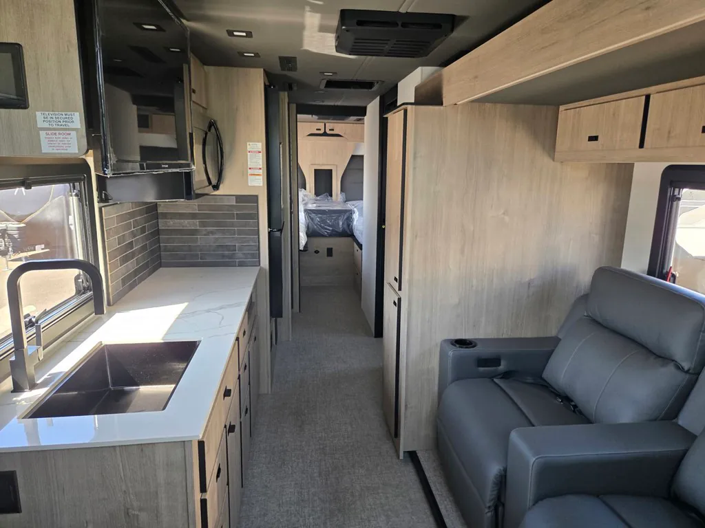 2027 Grand Design Lineage Series F 29AN5 — Class C motorhome kitchen with sink and counters beside a recliner, hallway, and visible rear bed