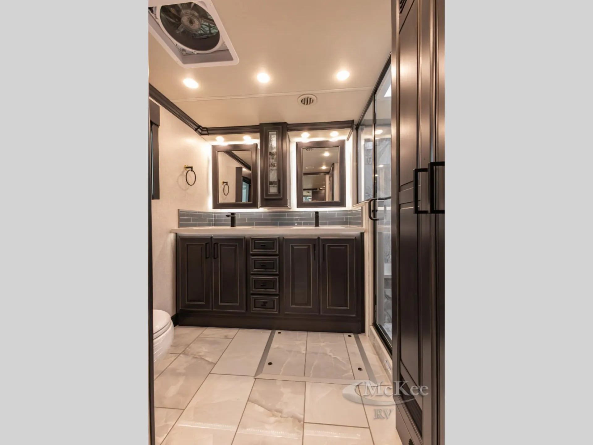 2027 Fleetwood Discovery LXE 40M — Bathroom vanity with double mirrors, wide countertop, backsplash, dark cabinetry, toilet edge, and glass shower enclosure