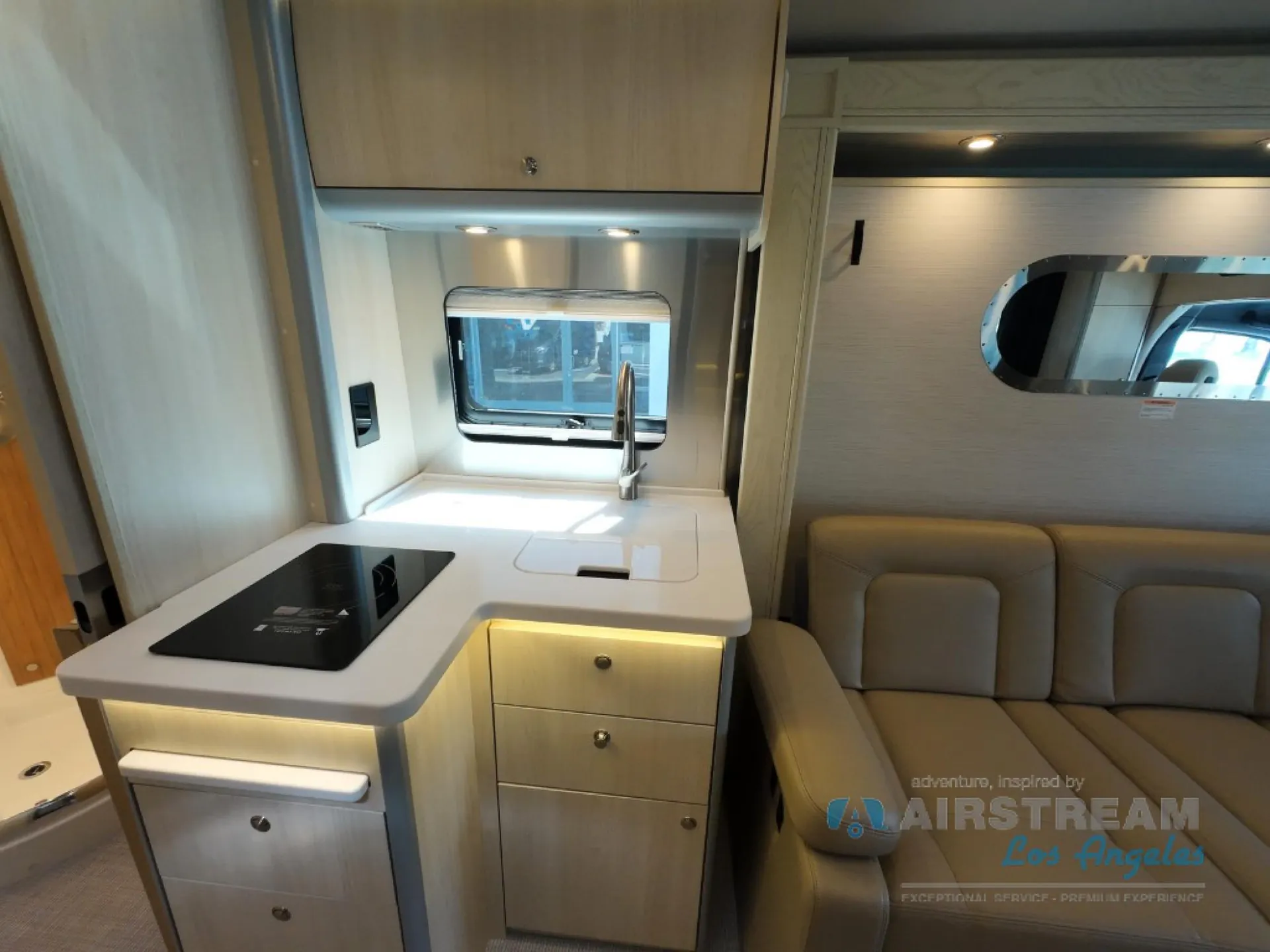 2026 Airstream Atlas Murphy Suite — Compact kitchen with induction cooktop, sink, drawers, and overhead cabinet beside a visible upholstered lounge seat