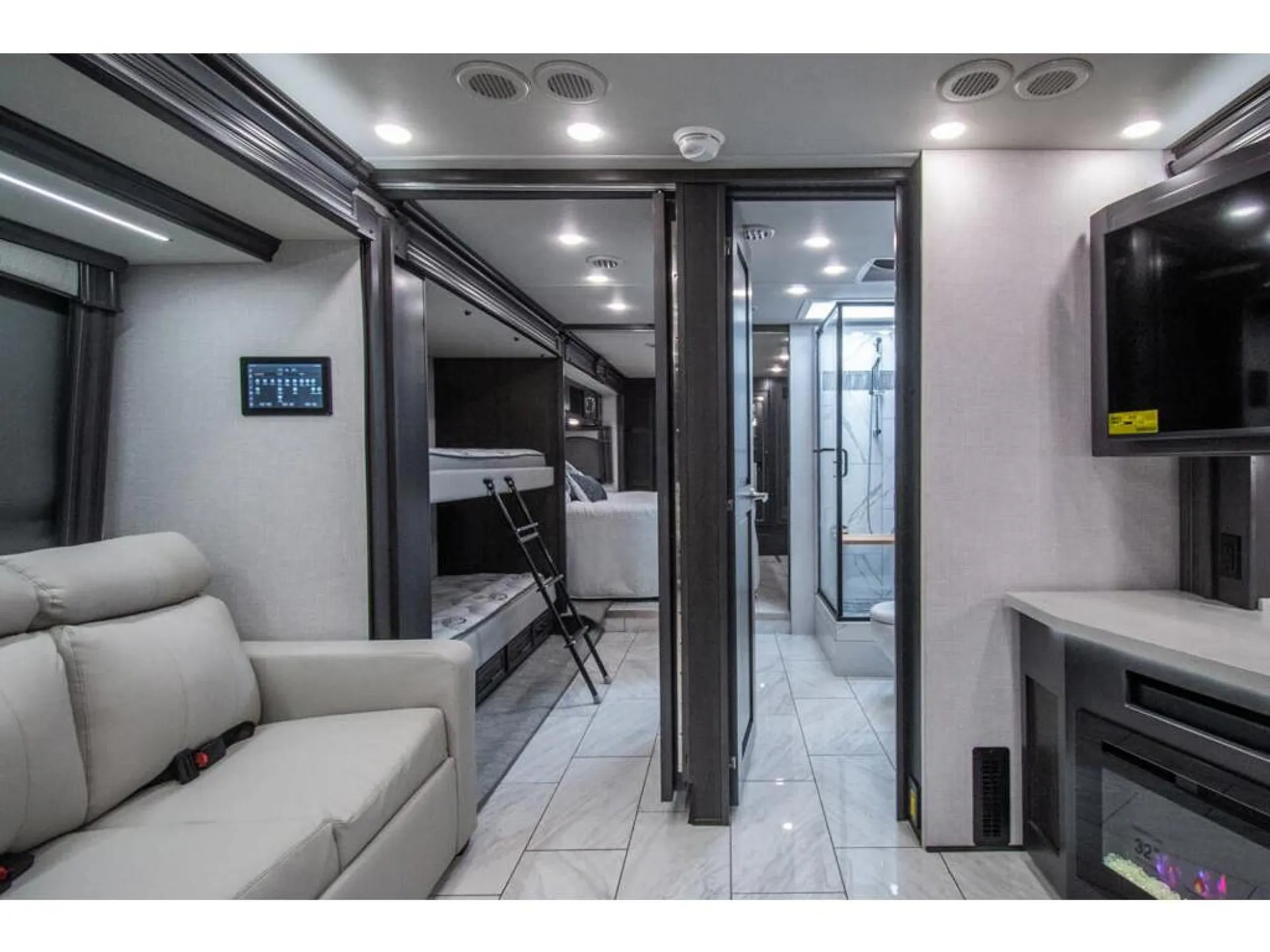 2026 Fleetwood Discovery LXE 40G — Lounge recliner, television and fireplace face a hallway leading past bunk beds toward a bathroom and rear sleeping area