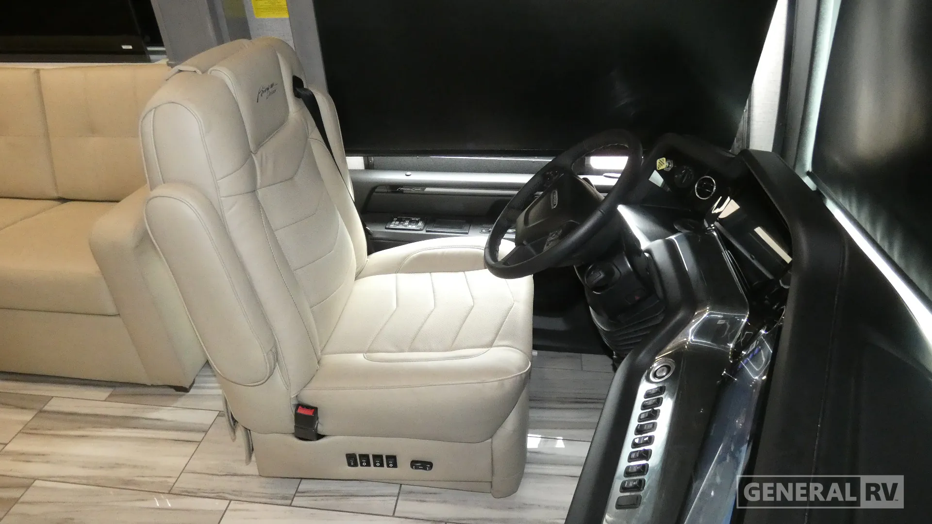 2026 American Coach American Dream 45A — Driver cockpit showing steering wheel, instrument controls, driver seat, dashboard, windshield, and adjacent lounge sofa