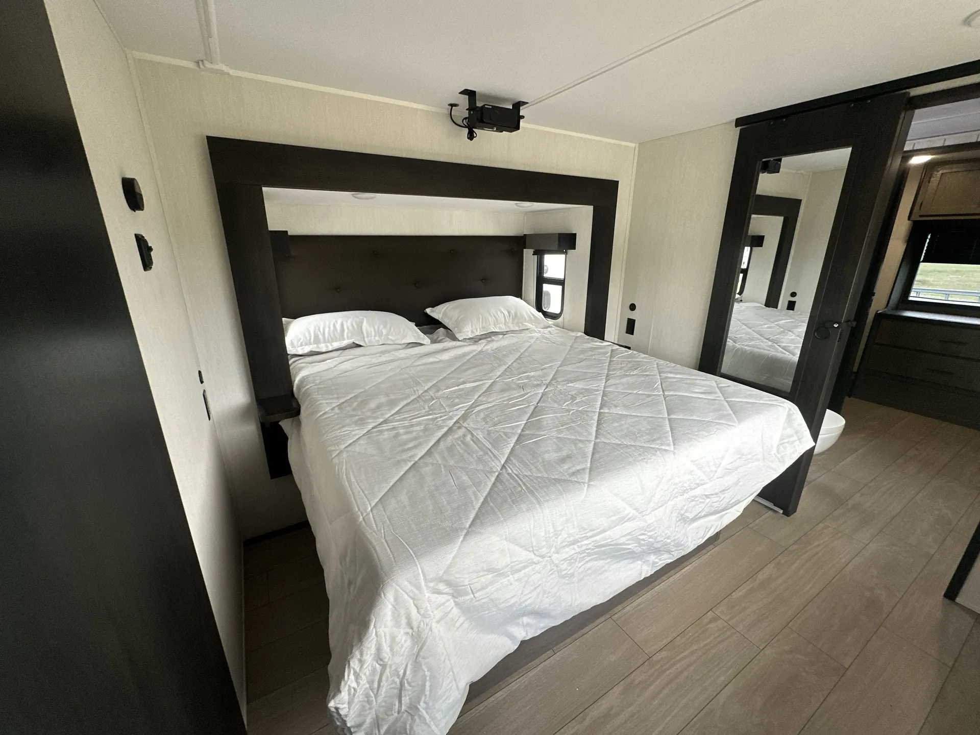 2026 Forest River Arctic Wolf 38LEAH.G — Main bedroom with a made bed, padded headboard, bedside surfaces, windows, and a wall-mounted projector