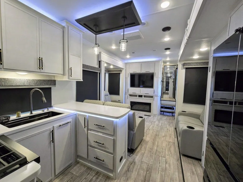 2025 Dutchmen Voltage 4225 — Fifth-wheel kitchen with sink, range, island, and cabinetry opening toward a living area with seating and television