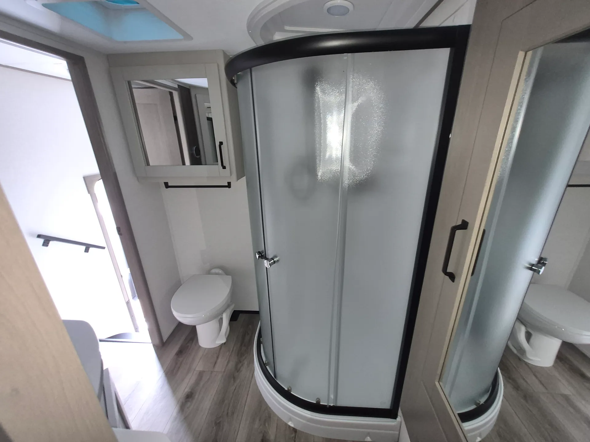 2027 Grand Design Reflection 362TBS — Bathroom with curved glass shower, toilet, mirrored cabinet, towel bar, and wood-look flooring