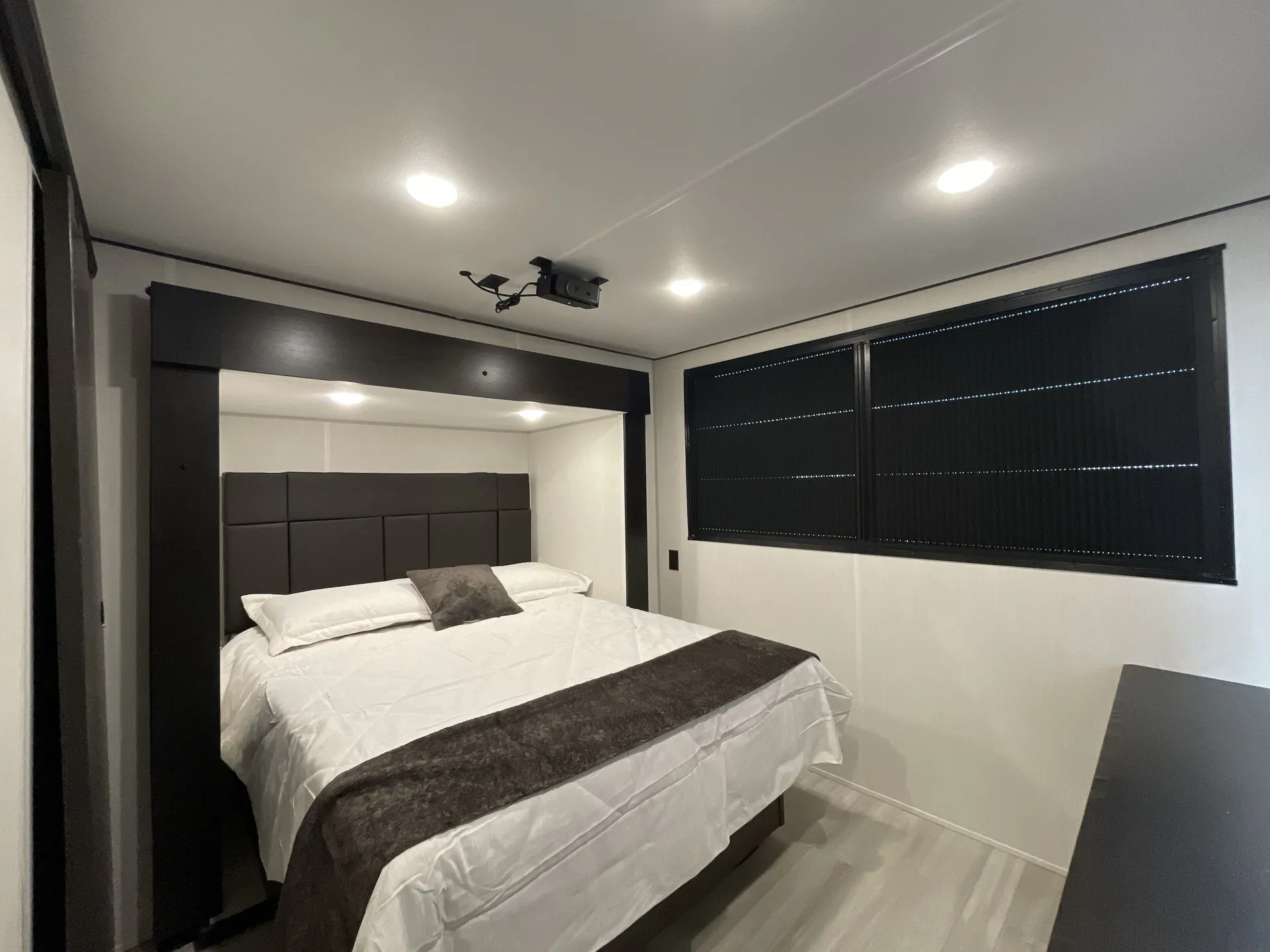 2026 Forest River Cherokee Wolf Pack 397STAY — Main bedroom with a made bed, padded headboard, wide window shades, ceiling-mounted projector, and dark overhead trim