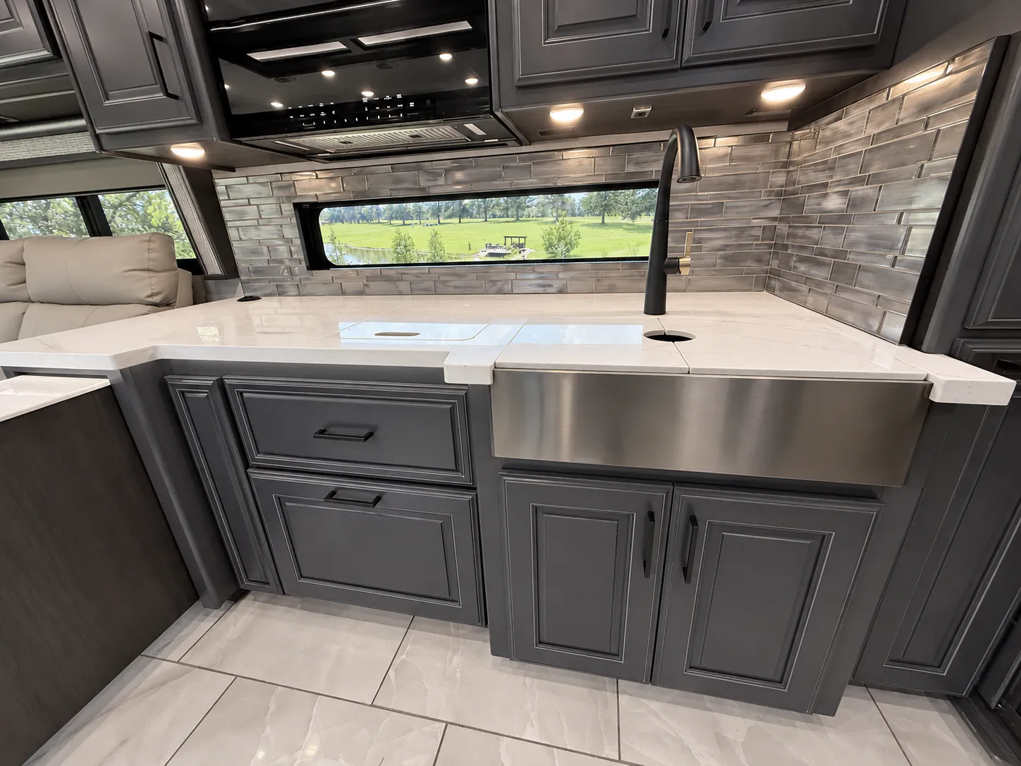2027 Fleetwood Discovery LXE 40M — Kitchen work area with large farmhouse sink, cooktop covers, white counter, tile backsplash, window, and overhead cabinetry