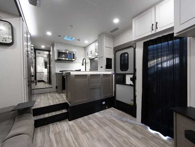 2025 Jayco Seismic Luxury Series 4113 — Fifth wheel kitchen with island sink, microwave, cabinetry, entry door, and adjacent reclining sofa