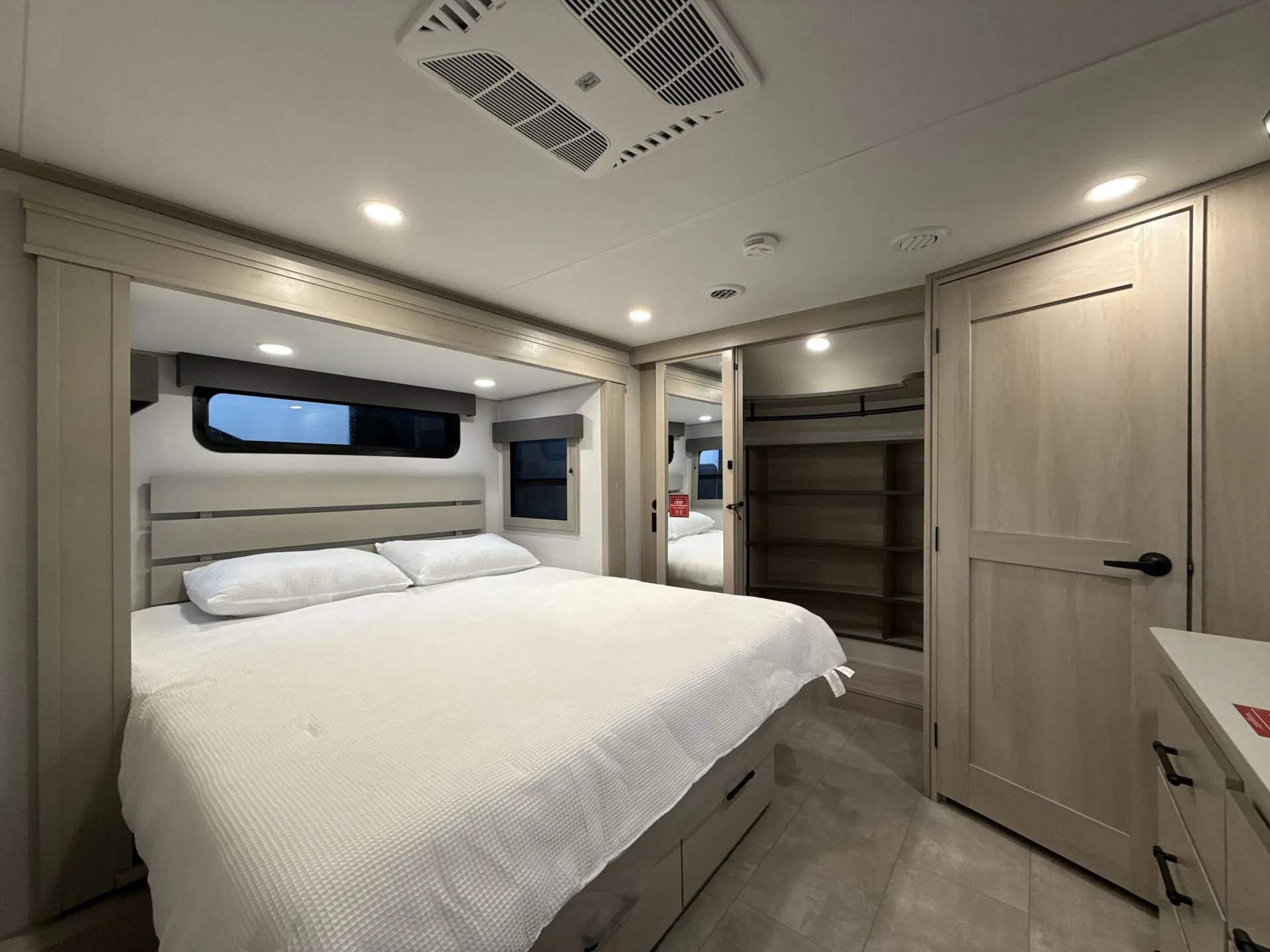2026 Grand Design Solitude 391DL — Main bedroom with a large bed, mirrored wardrobe, open shelving, dresser, windows, and overhead air-conditioning unit