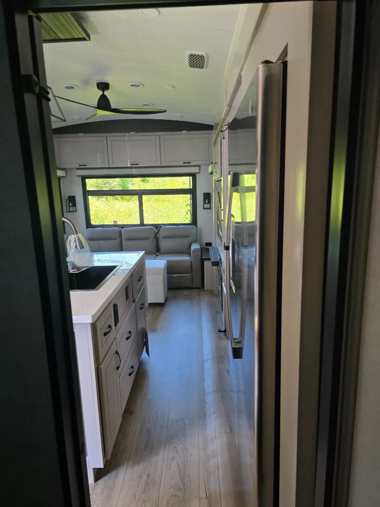 2026 Jayco North Point 377RLBH — Fifth-wheel interior overview showing a kitchen island, refrigerator, open aisle, ceiling fan, and rear sofa