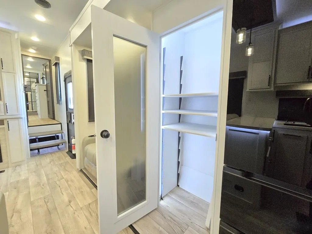 2025 Dutchmen Voltage 4225 — Fifth wheel kitchen cabinetry and refrigerator beside an open pantry, with a hallway and entry steps beyond