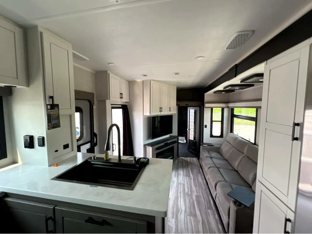 2025 Jayco Seismic Luxury Series 4113 — Fifth-wheel kitchen island with sink and cabinetry facing a recliner sofa and entertainment center