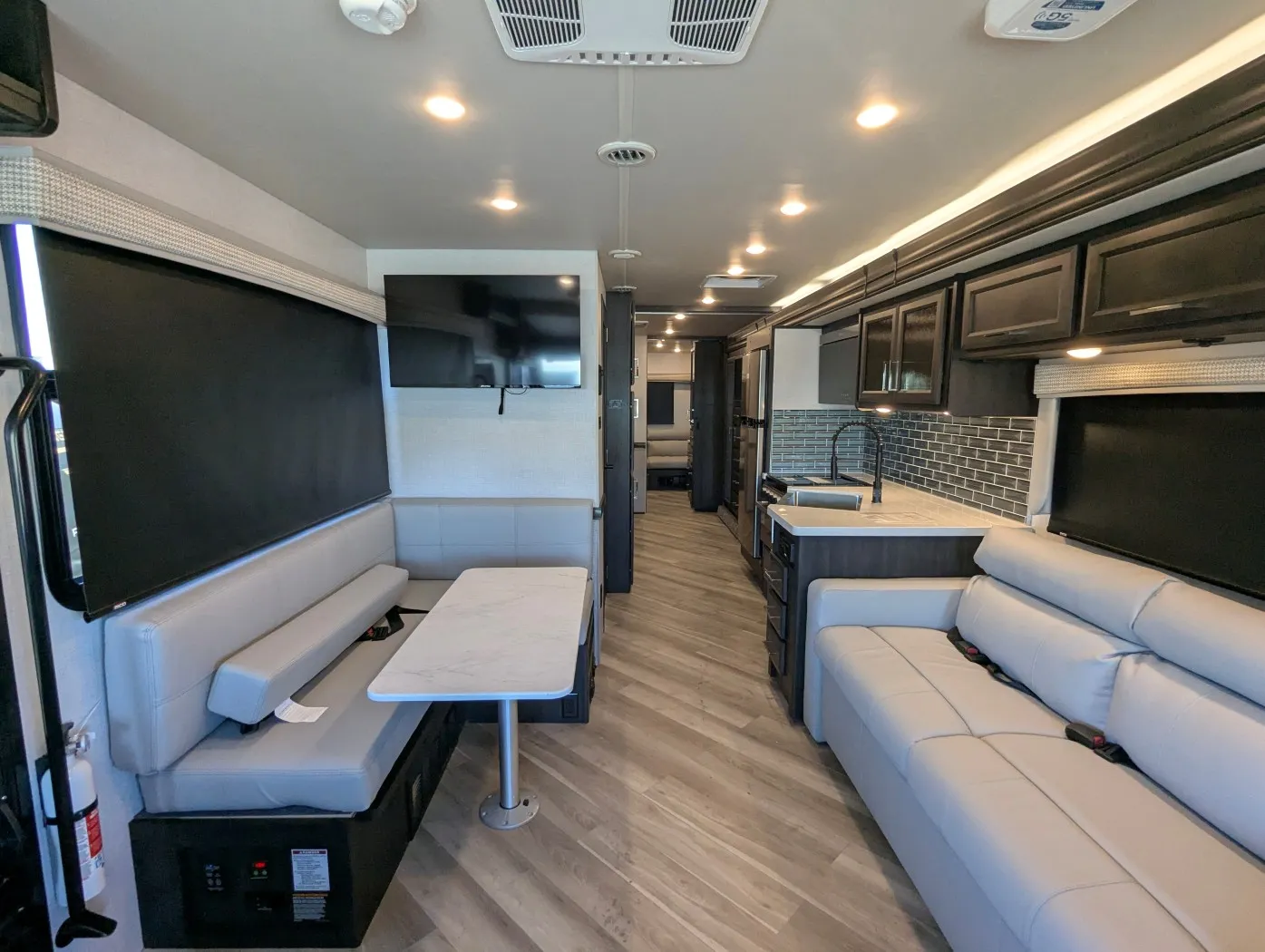 2026 Holiday Rambler Invicta 36Y — Dinette booth and table opposite a sofa, with television, sink, backsplash, and kitchen cabinetry visible along the aisle