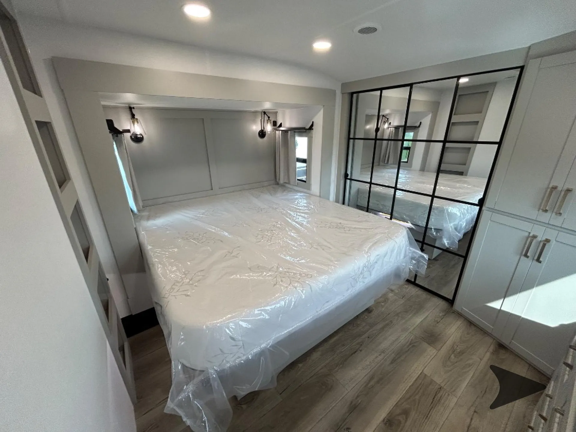 2026 Brinkley Model Z 3515 — Main bedroom with plastic-wrapped bed, mirrored wardrobe doors, bedside lights, windows, and wood-look flooring