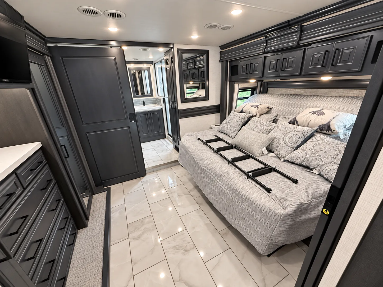2027 Fleetwood Discovery LXE 40M — Main bedroom with large bed, overhead cabinets, dresser, and a bathroom vanity visible through the rear doorway