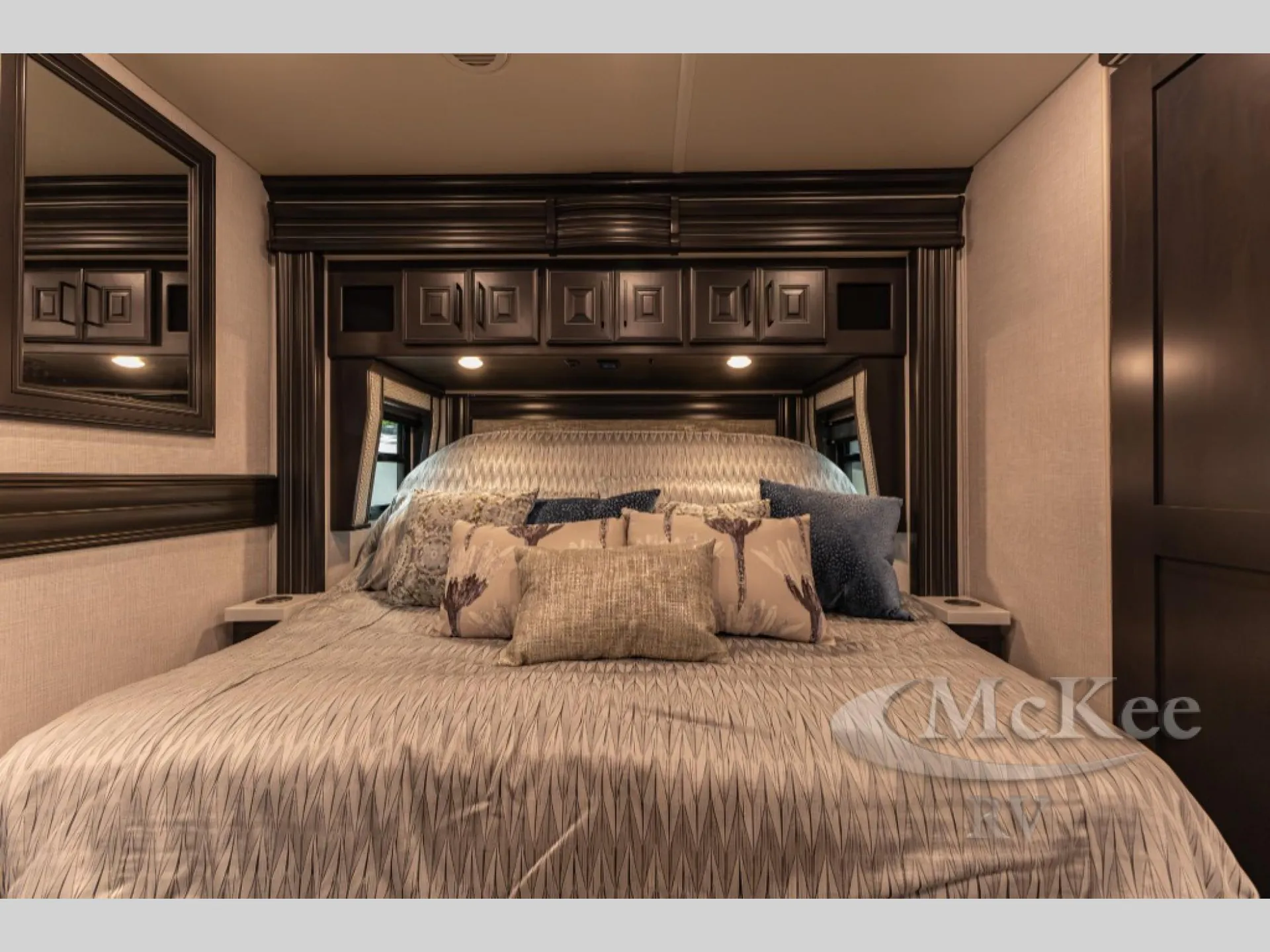 2027 Fleetwood Discovery LXE 40M — Front-facing view of the main bedroom bed beneath overhead cabinets, with bedside surfaces and surrounding cabinetry