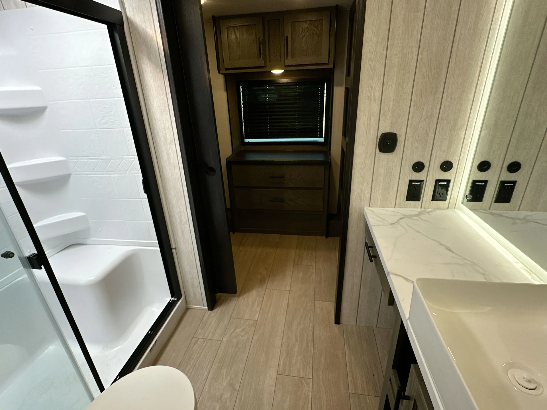 2026 Forest River Arctic Wolf 38LEAH.G — Fifth-wheel bathroom with a glass shower, toilet edge, vanity, and adjacent dresser visible beyond the doorway
