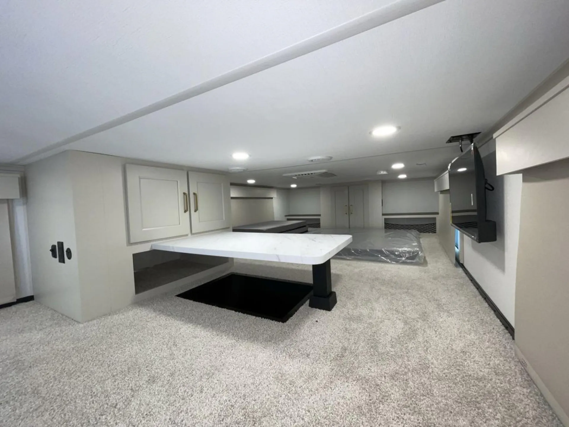 2026 Grand Design Foundation 42GD — Spacious carpeted destination trailer loft with central table, built-in cabinets, mattresses, and wall-mounted television