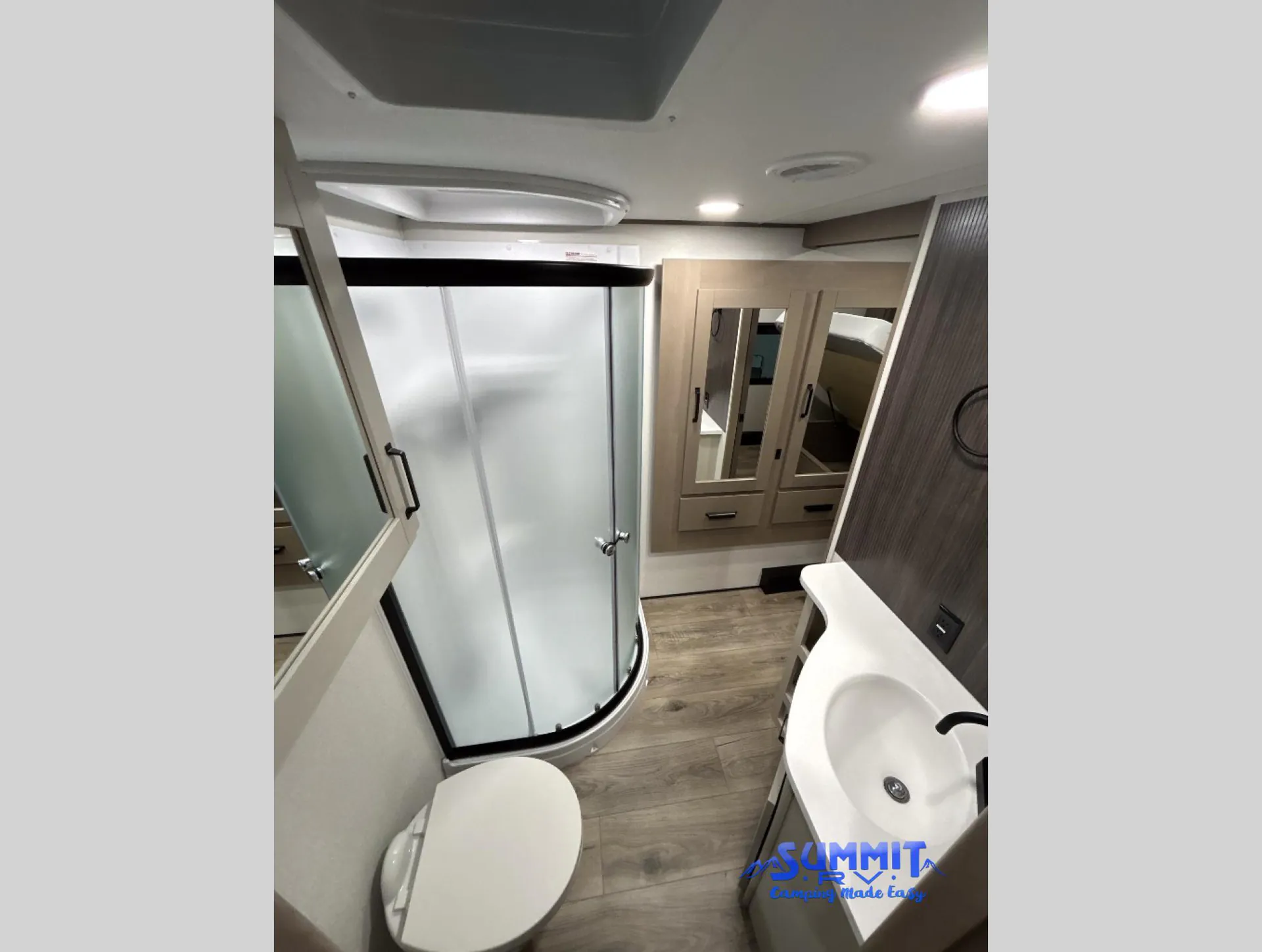 2027 Grand Design Reflection 362TBS — Fifth-wheel bathroom with curved shower, toilet, vanity sink, mirrored cabinets, and wood-look flooring