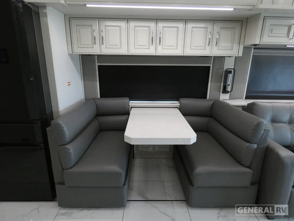 2026 American Coach American Dream 45A — U-shaped dining booth and table beside a gray sofa, with overhead cabinets and window shades
