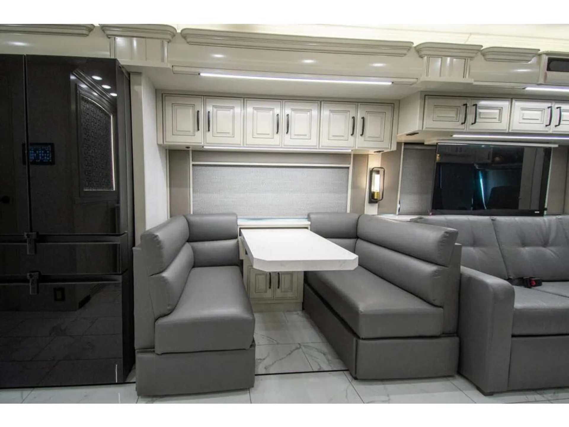 2026 American Coach American Dream 45A — Motorhome dinette with opposing benches and table beside a refrigerator, with adjacent living sofa and entertainment wall visible