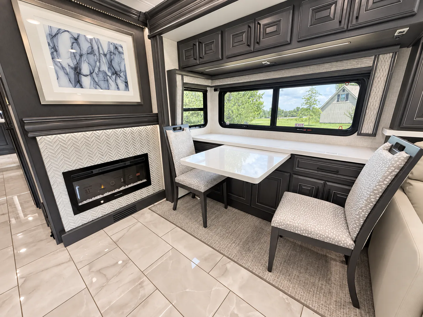 2027 Fleetwood Discovery LXE 40M — Freestanding dining table with two upholstered chairs, window-side counter, fireplace, and overhead cabinets