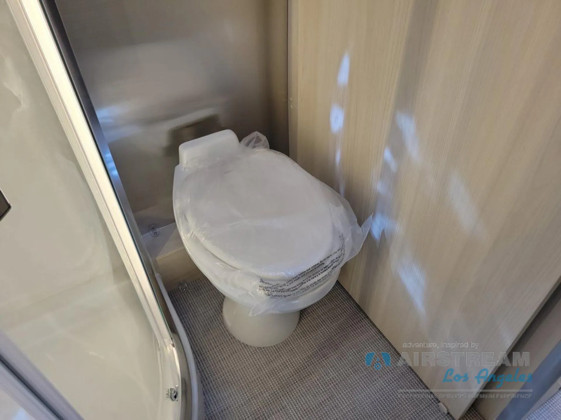 2026 Airstream Atlas Murphy Suite — Compact bathroom toilet with protective wrapping, gray floor mat, and surrounding wood cabinetry
