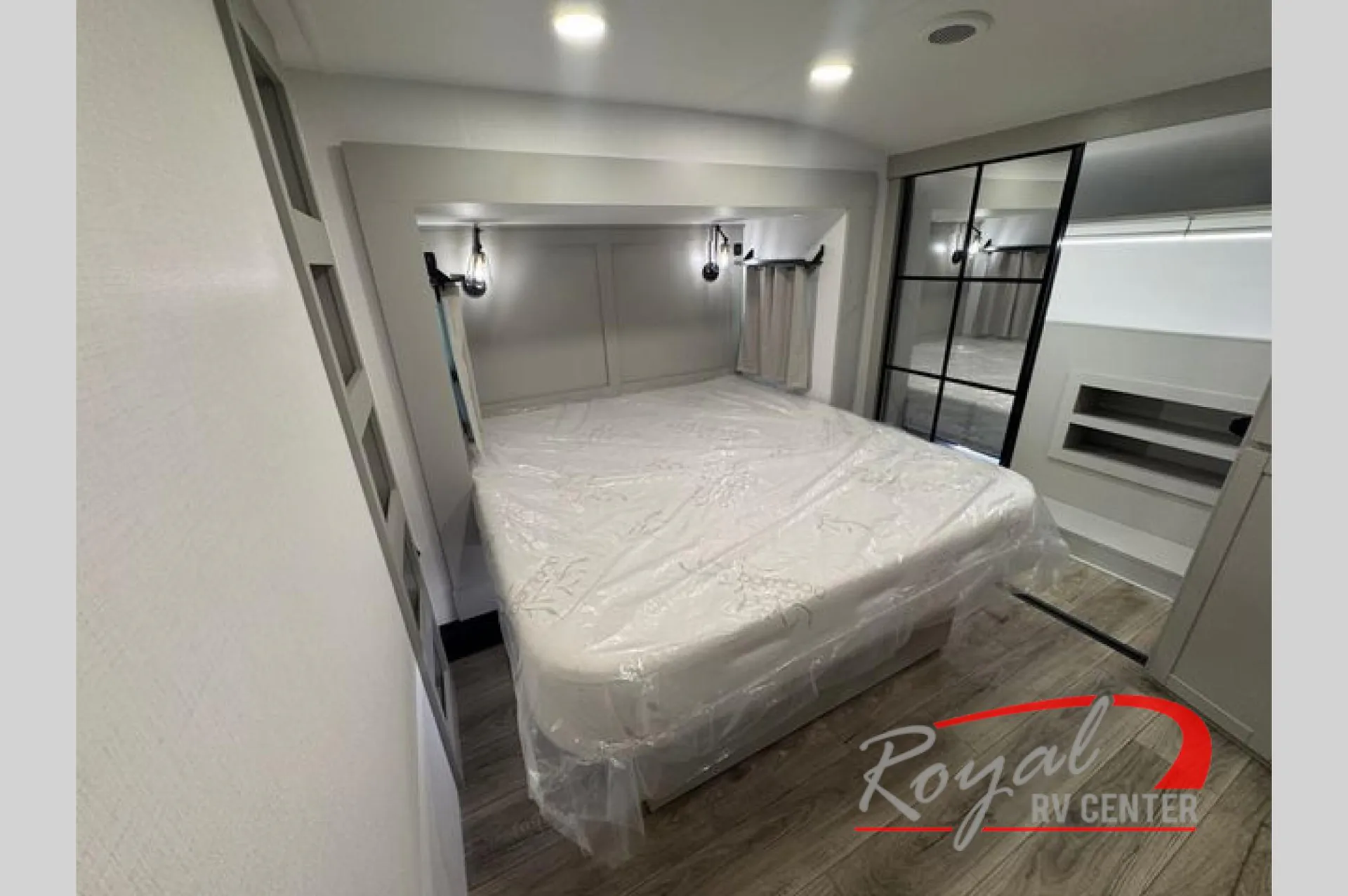 2026 Brinkley Model Z 3610 — Fifth-wheel bedroom with a plastic-wrapped bed, mirrored wardrobe doors, bedside lighting, and wall shelving