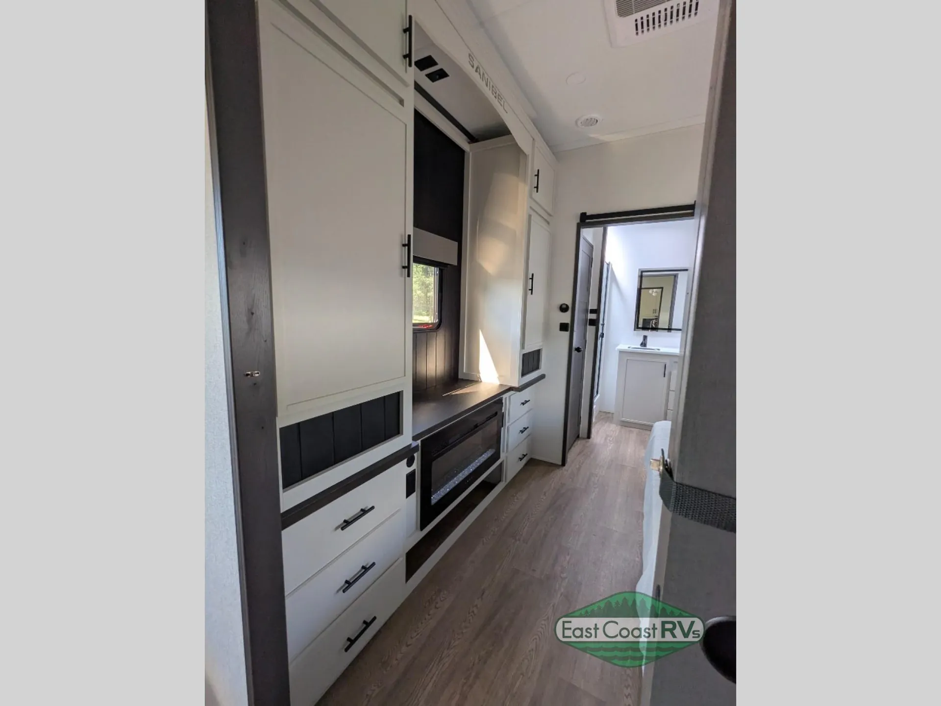 2027 Forest River Sanibel 40BUNK — Living entertainment wall with fireplace and storage cabinets, with a bathroom vanity visible down the hall