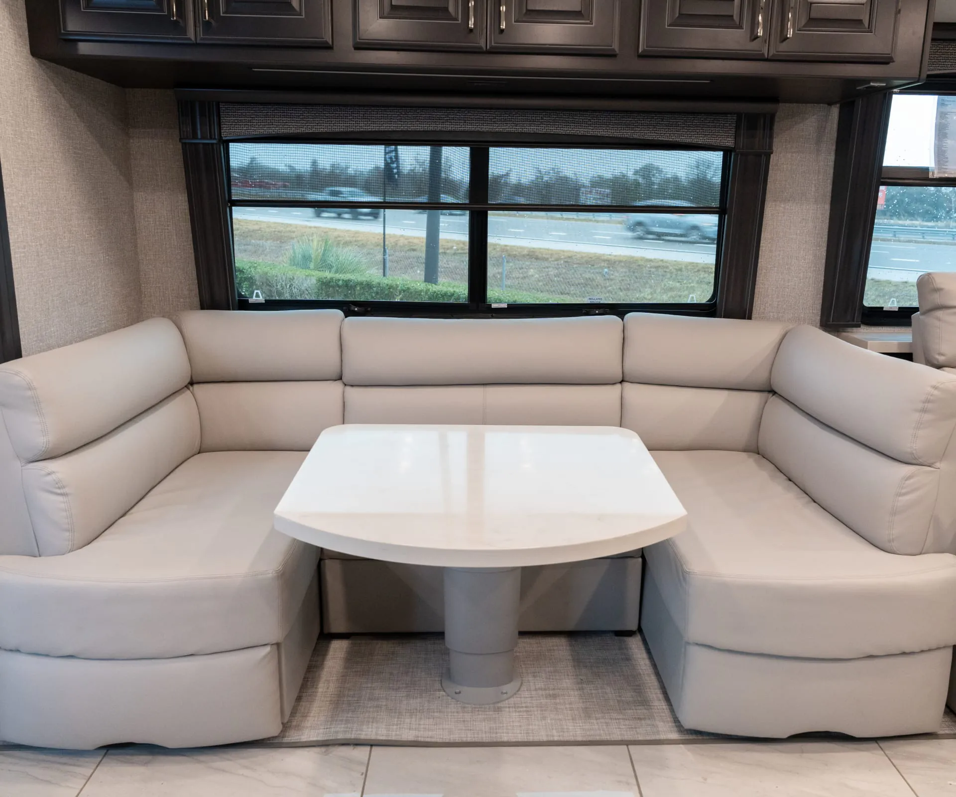 2026 Fleetwood Discovery LXE 40G — U-shaped lounge seating surrounding a freestanding table beneath large windows and overhead cabinets