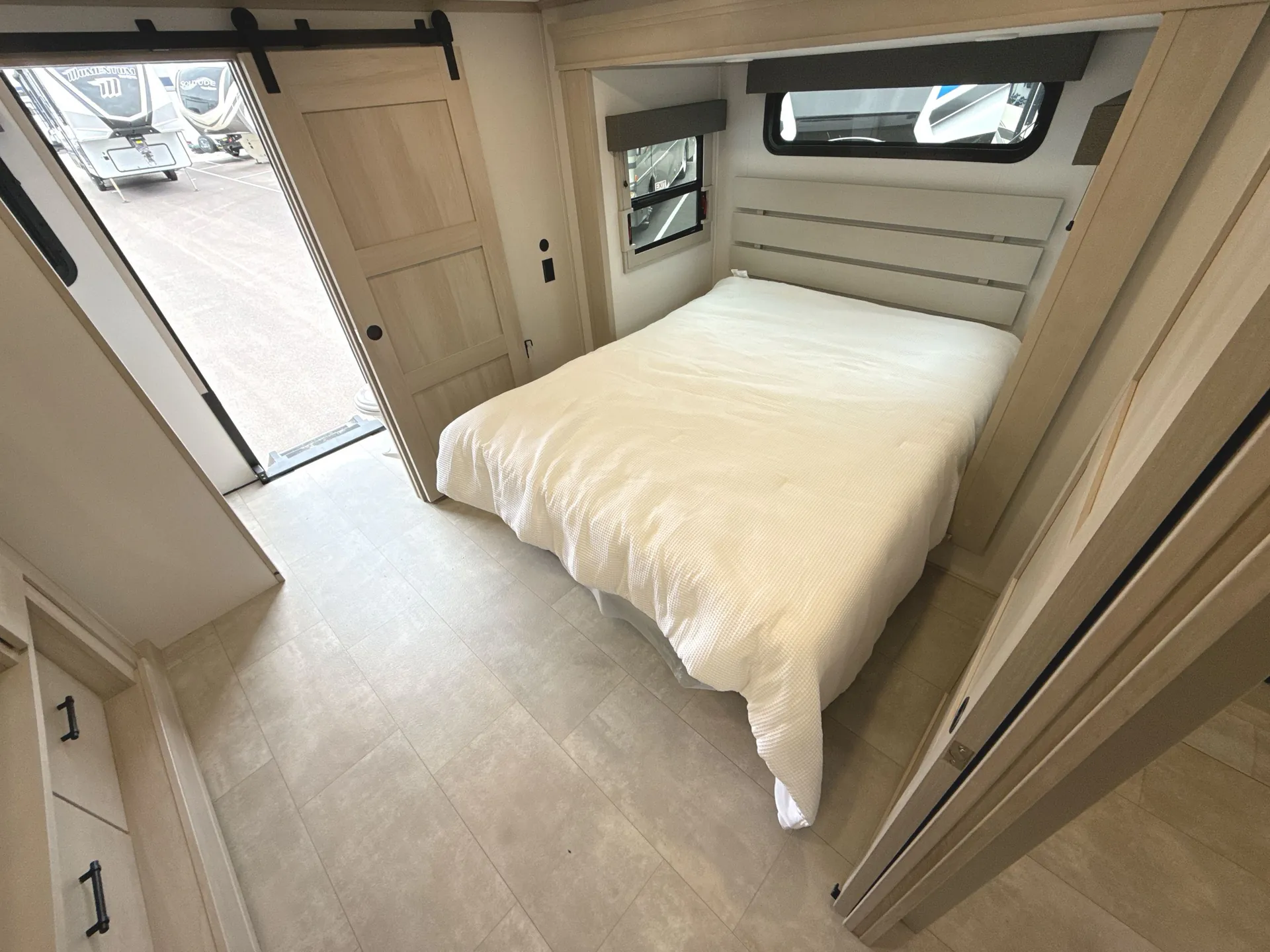 2026 Grand Design Solitude 391DL — Main bedroom with a made bed, surrounding cabinetry, windows, sliding door, and open entry to the trailer