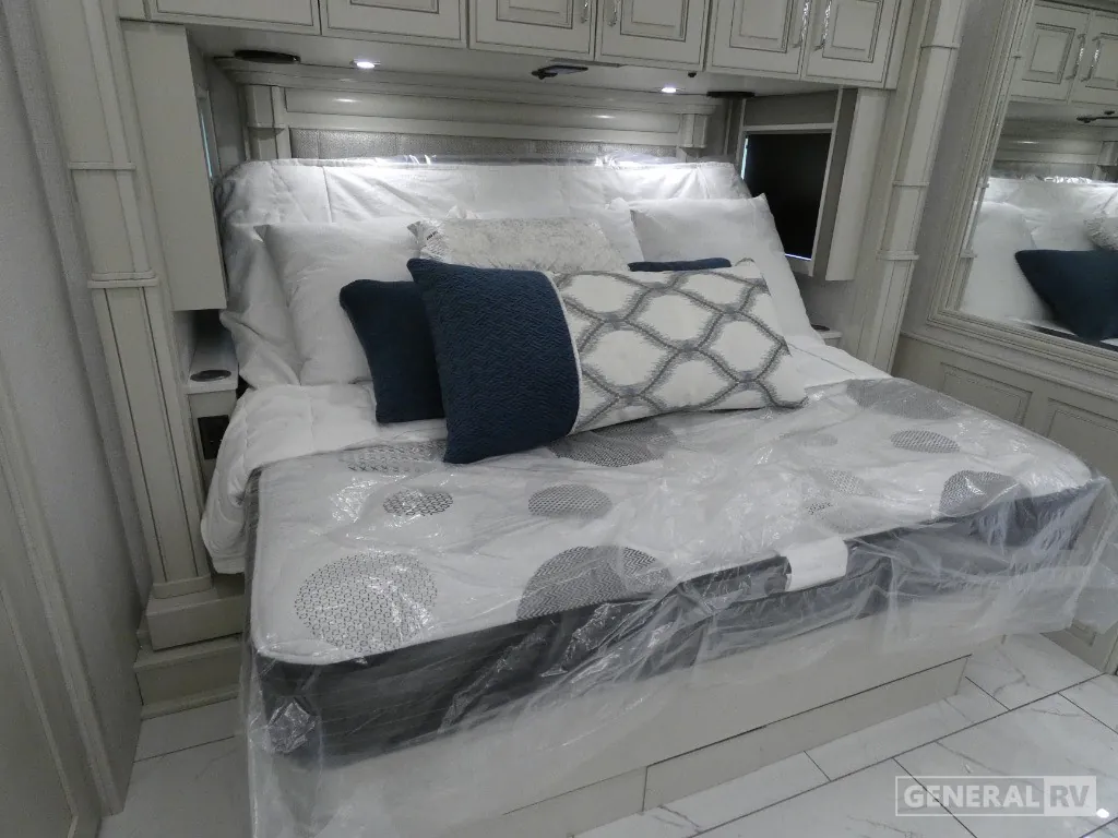 2026 American Coach American Dream 45A — Main bedroom bed with layered pillows, overhead cabinets, bedside storage, and a mirrored wardrobe panel