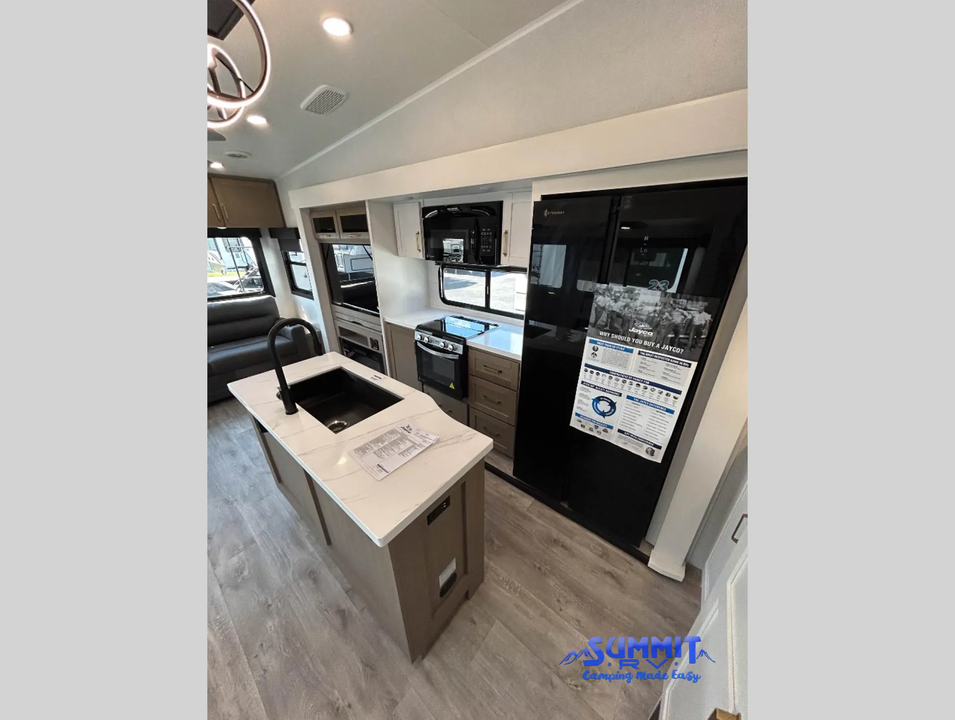 2027 Jayco Eagle 355MBQS — Fifth wheel kitchen with island sink, range, microwave, refrigerator, and sofa visible beyond the counters