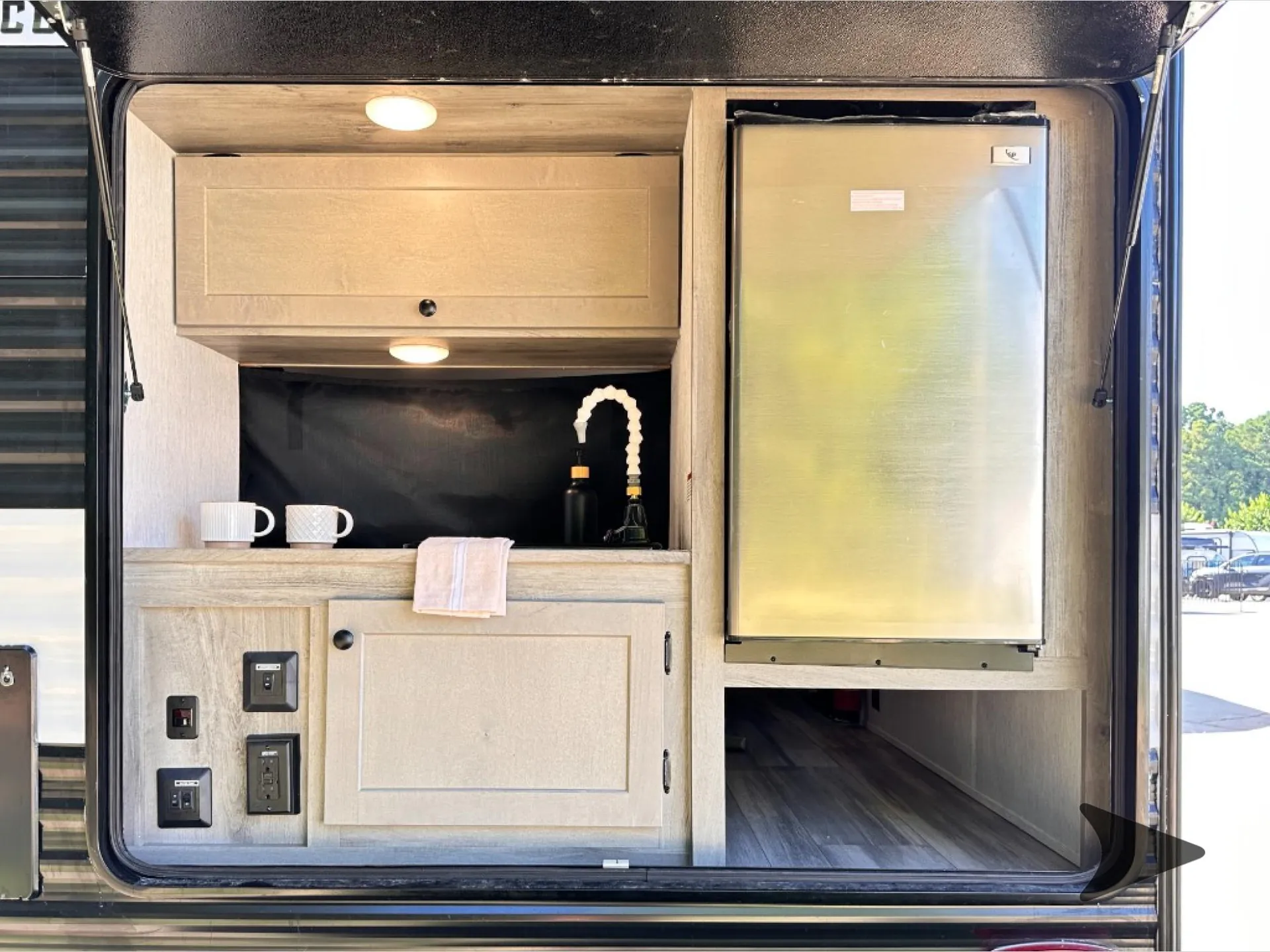 2026 Wayfinder Go Lite Solo — Close photograph through the exterior hatch showing the installed sink, faucet, counter, cabinetry, and refrigerator
