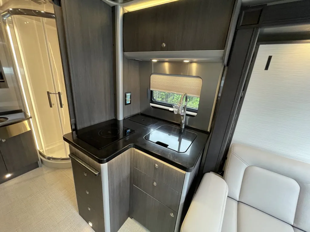 2026 Airstream Atlas Murphy Suite — Class B van kitchen with sink, induction cooktop, cabinets, adjacent lounge seat, and bathroom doorway