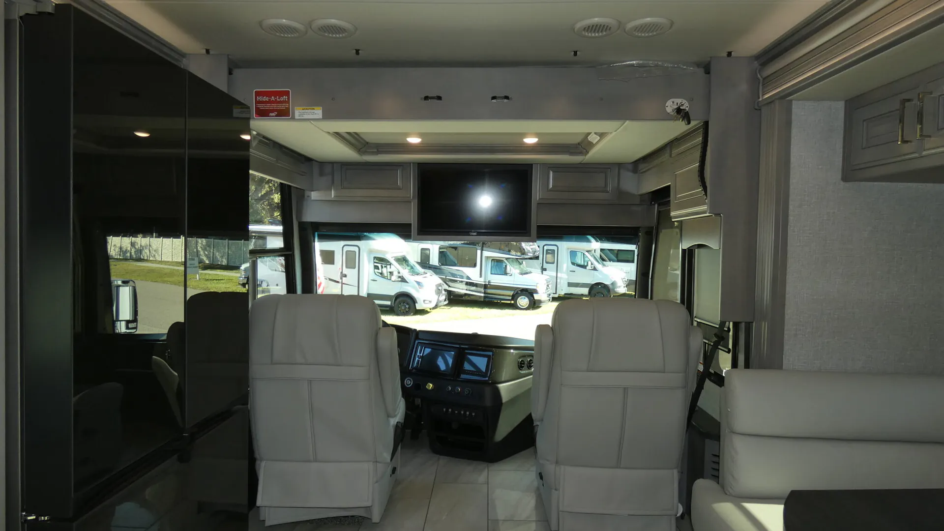 2026 Fleetwood Discovery LXE 40G — Class A cockpit with driver and passenger seats, dashboard controls, overhead television, and adjacent lounge seating