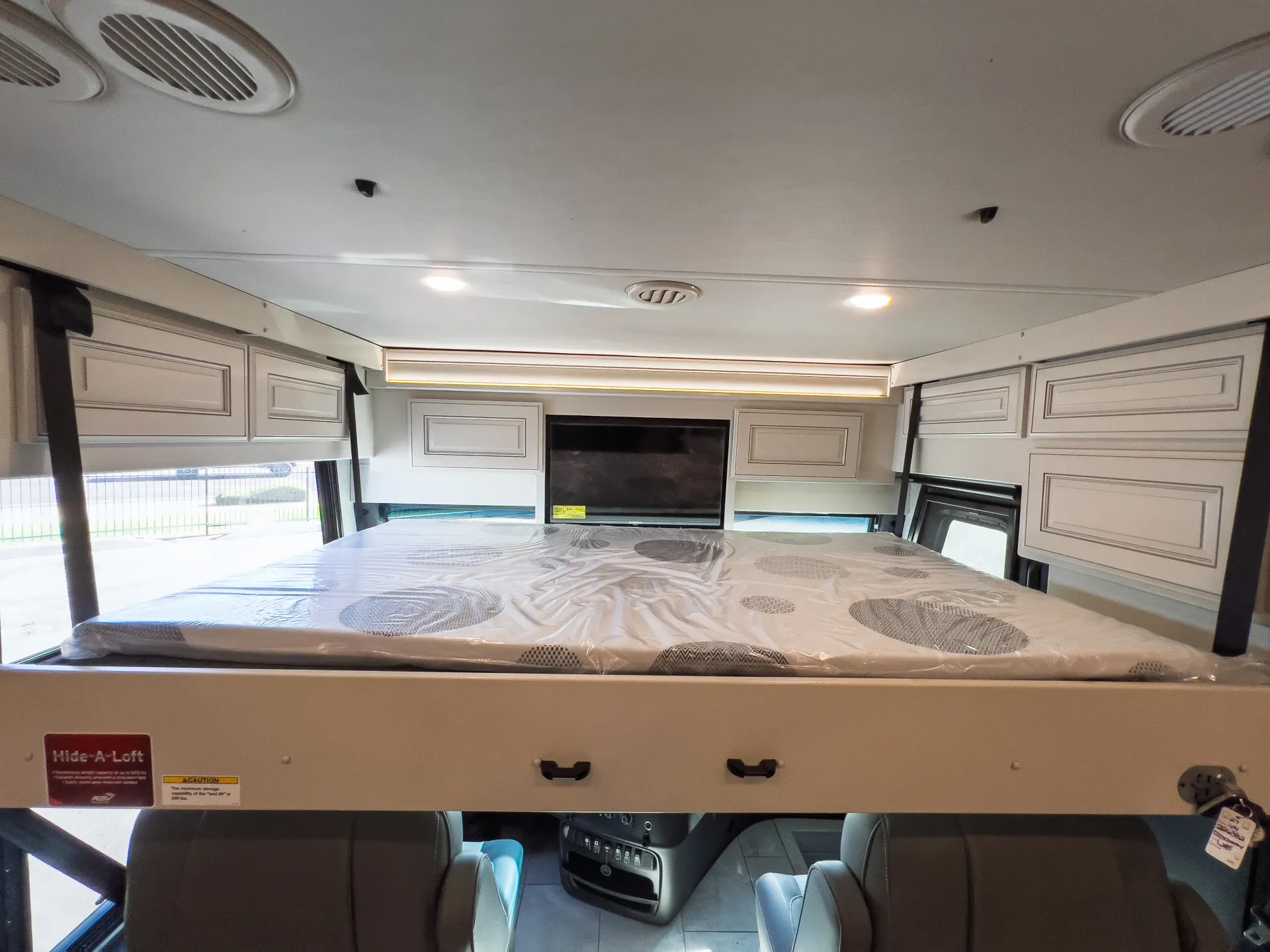 2027 Fleetwood Discovery LXE 40M — Over-cab loft bed with mattress, surrounding storage cabinets, television, and cockpit visible below