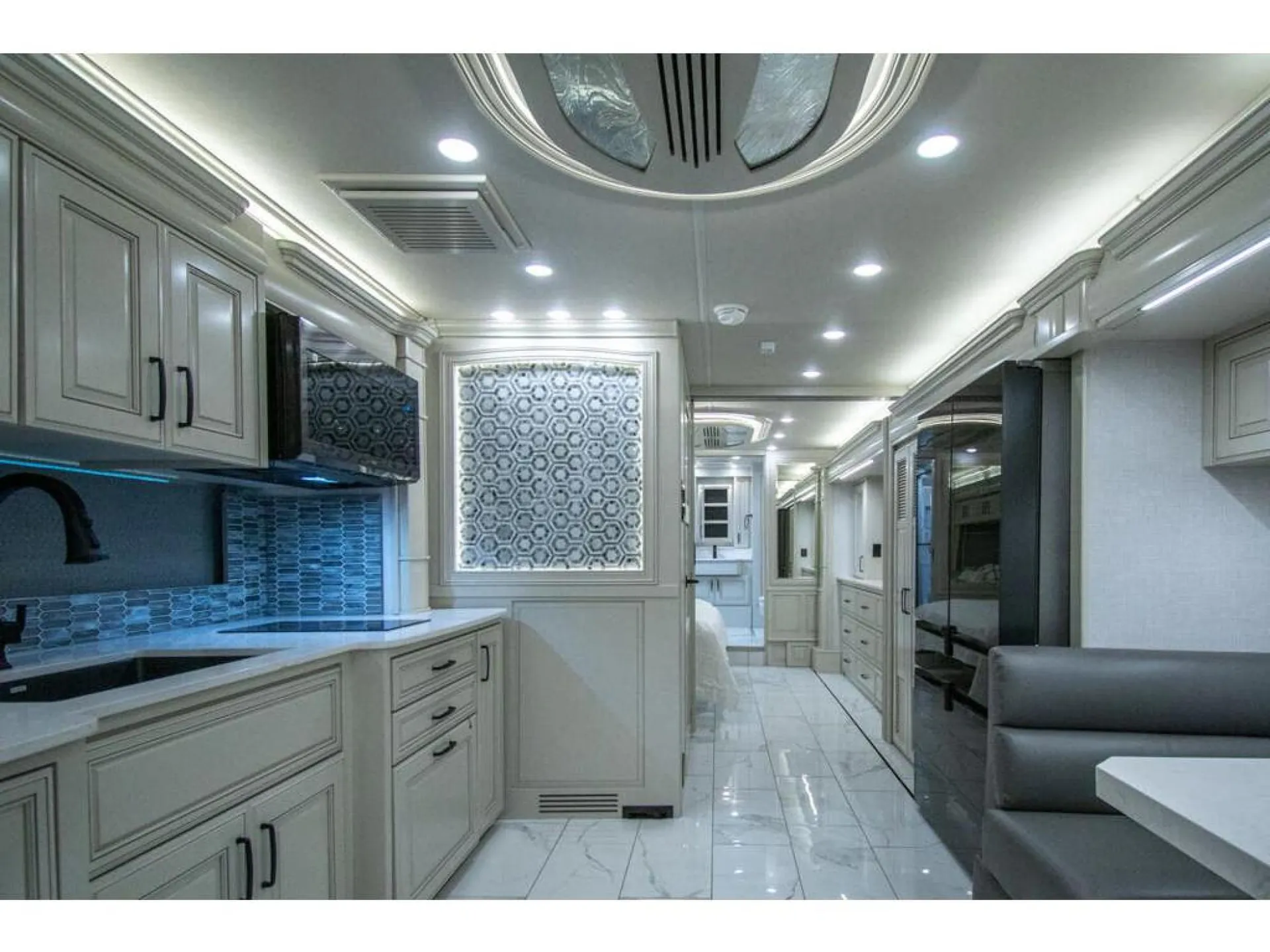 2026 American Coach American Dream 45A — Kitchen with sink, blue backsplash, cabinetry, and refrigerator beside a corridor leading toward seating and a bed
