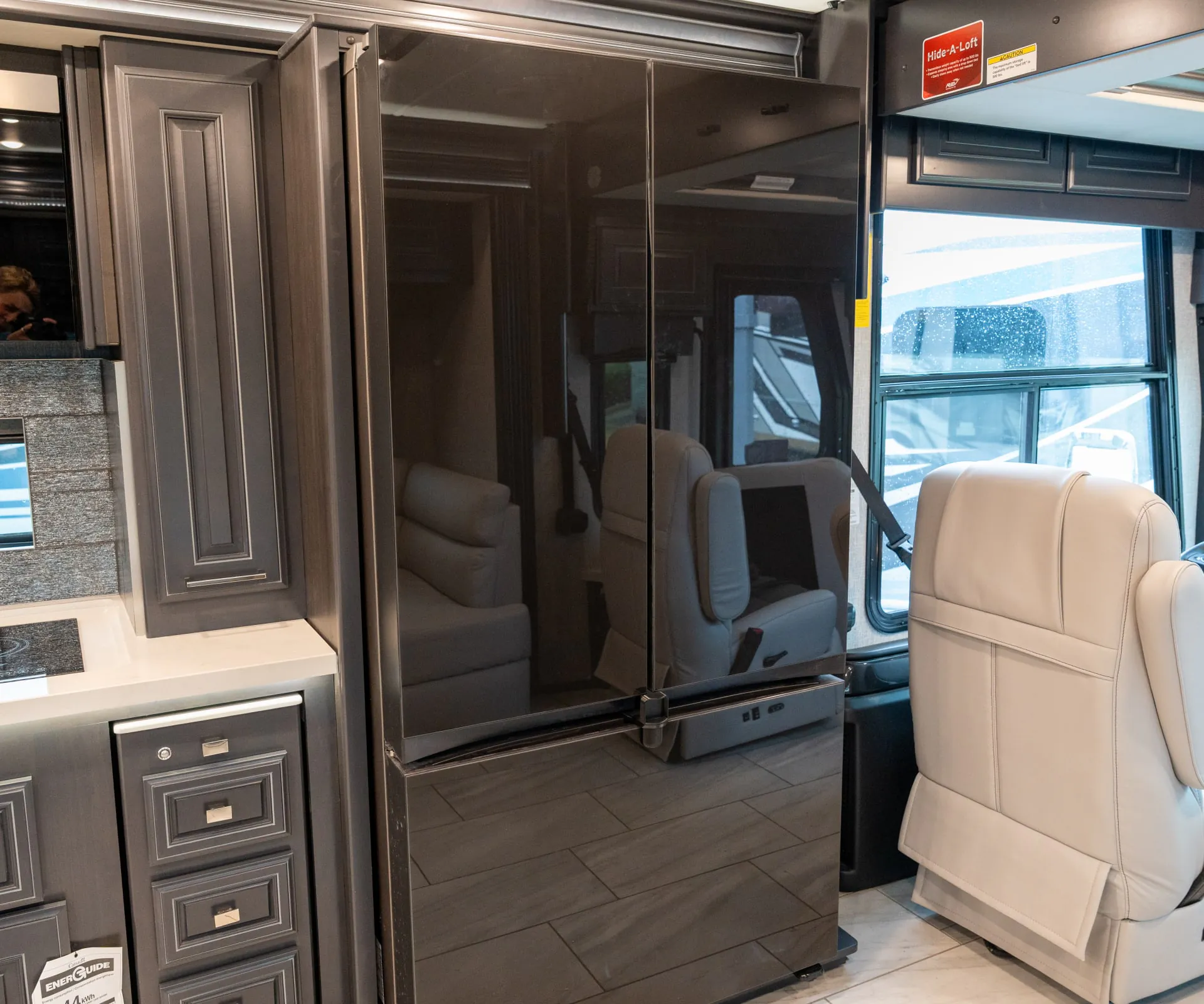 2026 Fleetwood Discovery LXE 40G — Tall refrigerator beside kitchen cabinetry, with a captain’s chair and front cab window visible in the cockpit