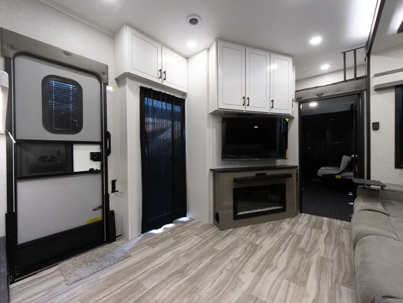 2025 Jayco Seismic Luxury Series 4113 — Living area entertainment center with television, fireplace, storage cabinets, entry door, and sofa edge