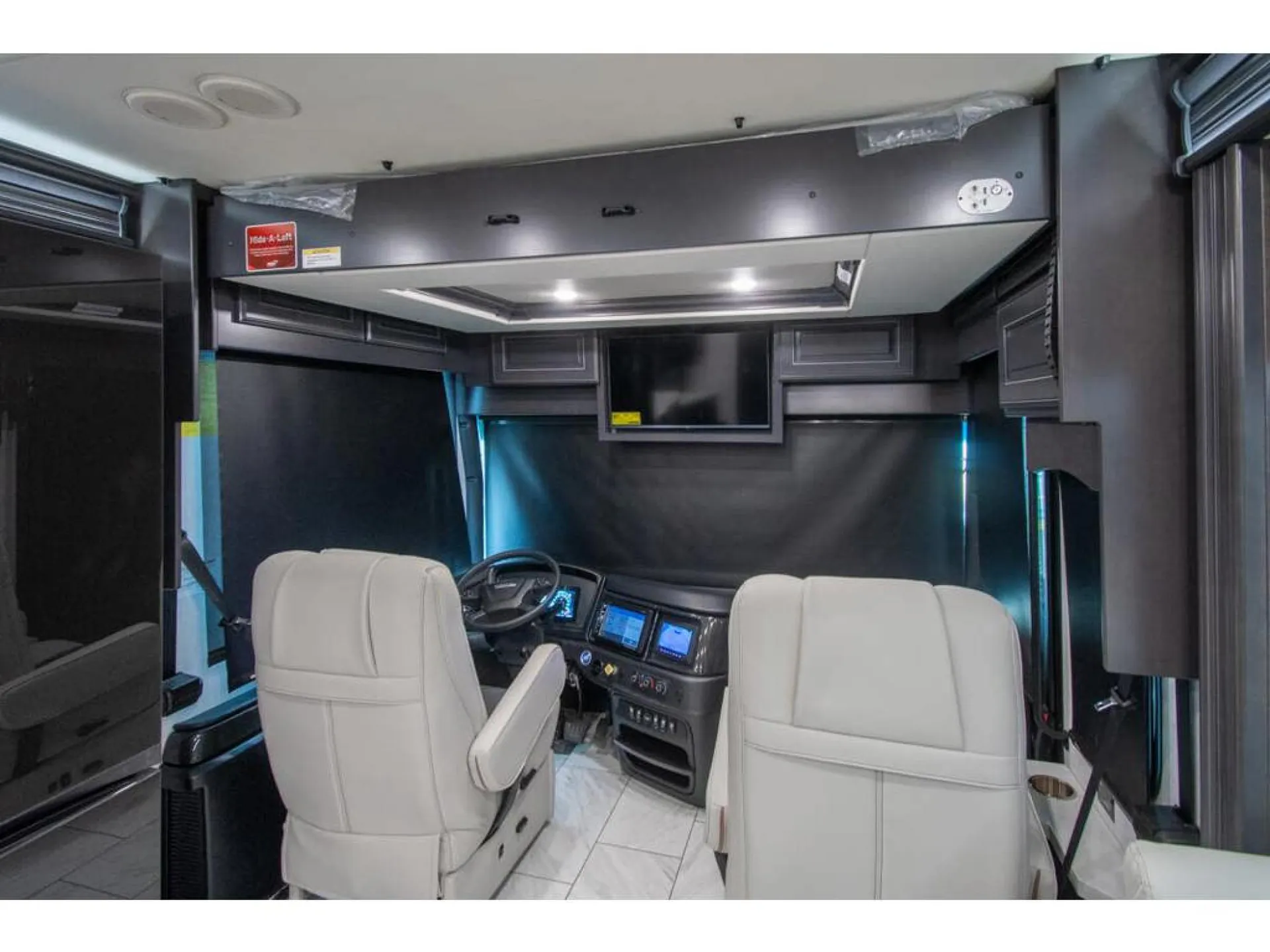 2026 Fleetwood Discovery LXE 40G — Driver cockpit with steering wheel, illuminated instrument panel, control screens, captain’s chairs, and overhead television
