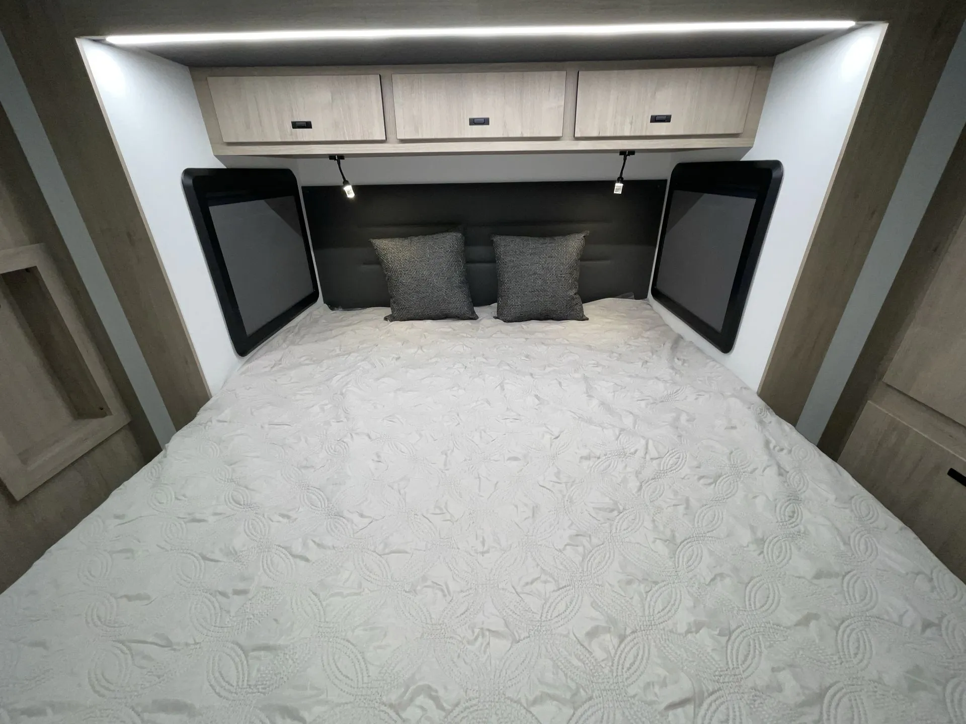 2027 Grand Design Lineage Series F 31ZW — Main bedroom bed beneath overhead cabinets, flanked by windows and illuminated reading lights