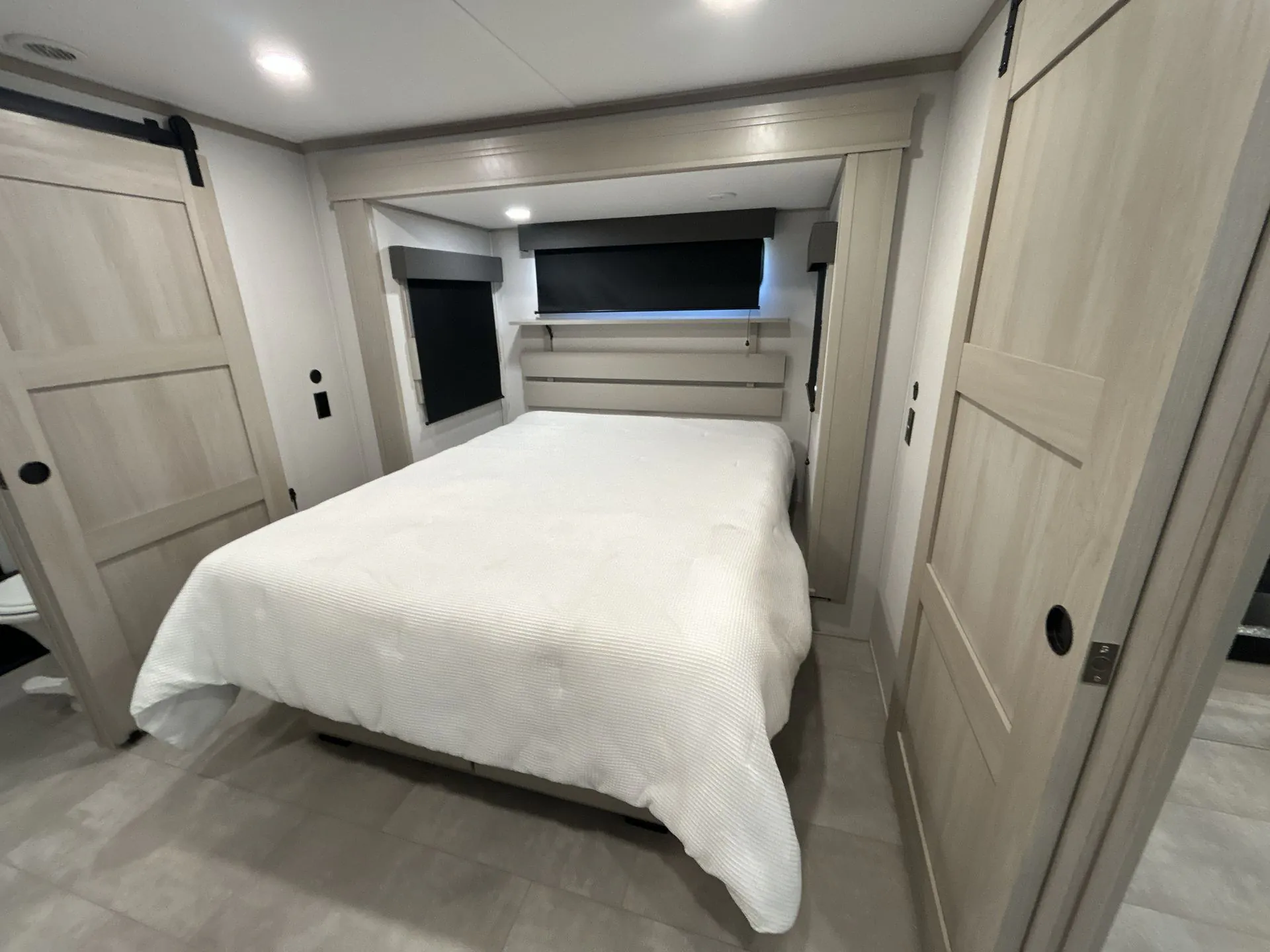 2026 Grand Design Solitude 391DL — Main bedroom with a made bed, blackout shades, side wardrobes, and light wood cabinetry