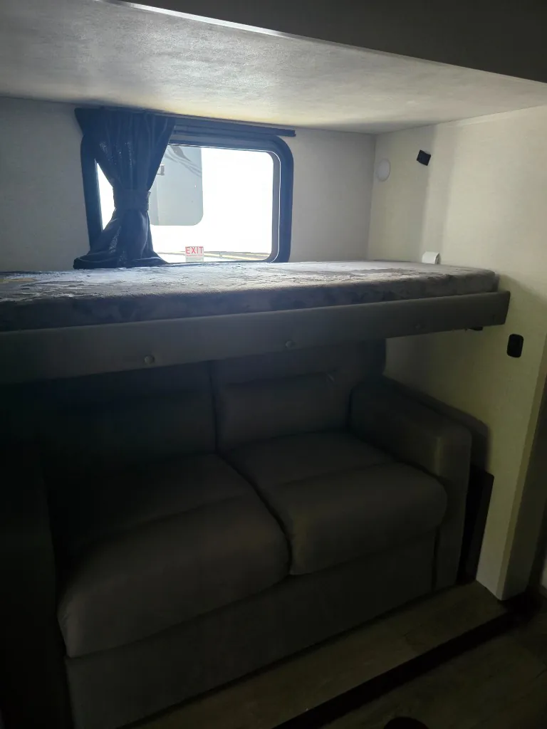 2026 Jayco North Point 377RLBH — Over-cab loft bunk above a compact sofa, with a curtained window and low ceiling