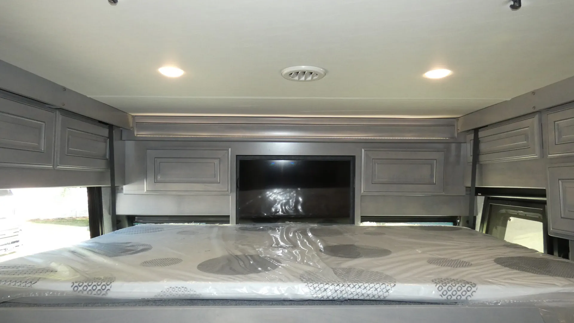2026 Fleetwood Discovery LXE 40G — Over-cab loft bed with plastic-wrapped mattress, surrounding cabinets, and a wall-mounted television