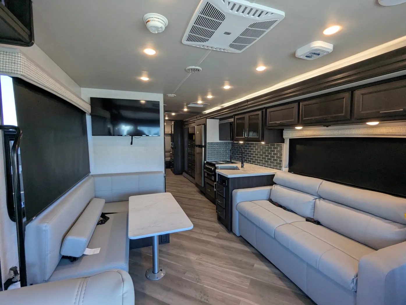 2026 Holiday Rambler Invicta 36Y — Living area with sofa and television beside a dinette, with the kitchen island and cabinetry extending down the aisle