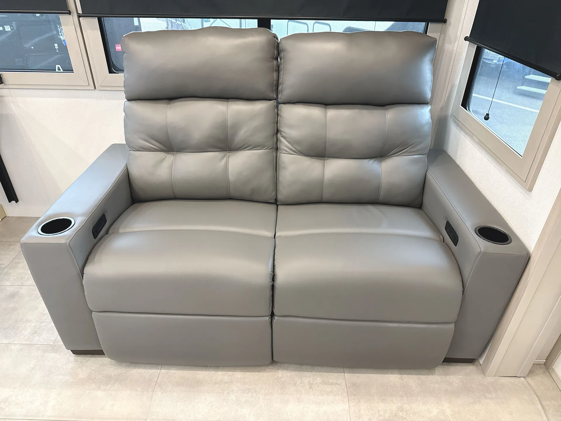 2026 Grand Design Solitude 391DL — Living area with a wall-mounted reclining loveseat, integrated cupholders, windows, and roller shades