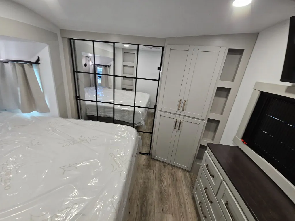 2026 Brinkley Model Z 3515 — Main bedroom with wrapped bed, black-framed glass divider, wardrobe cabinets, dresser, and television