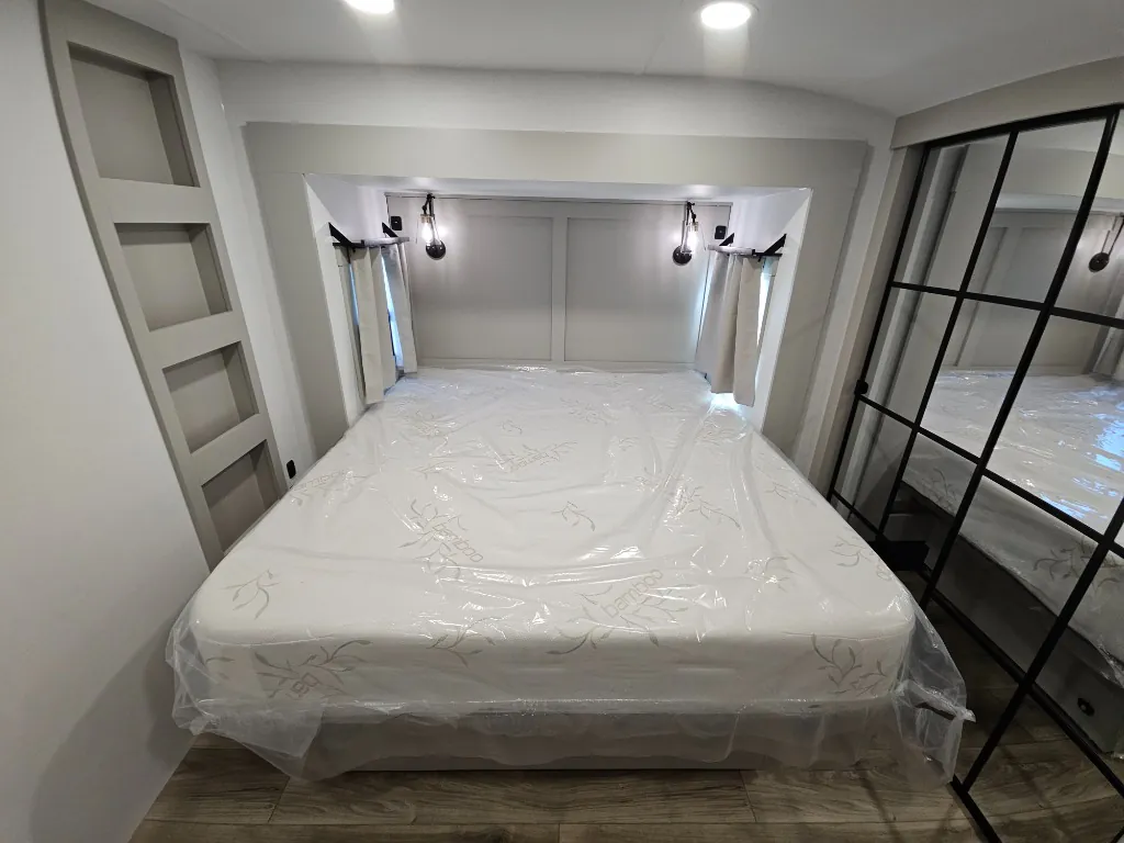 2026 Brinkley Model Z 3515 — Fifth wheel bedroom centered on a covered main bed with bedside reading lights, shelves, curtains, and mirrored wardrobe