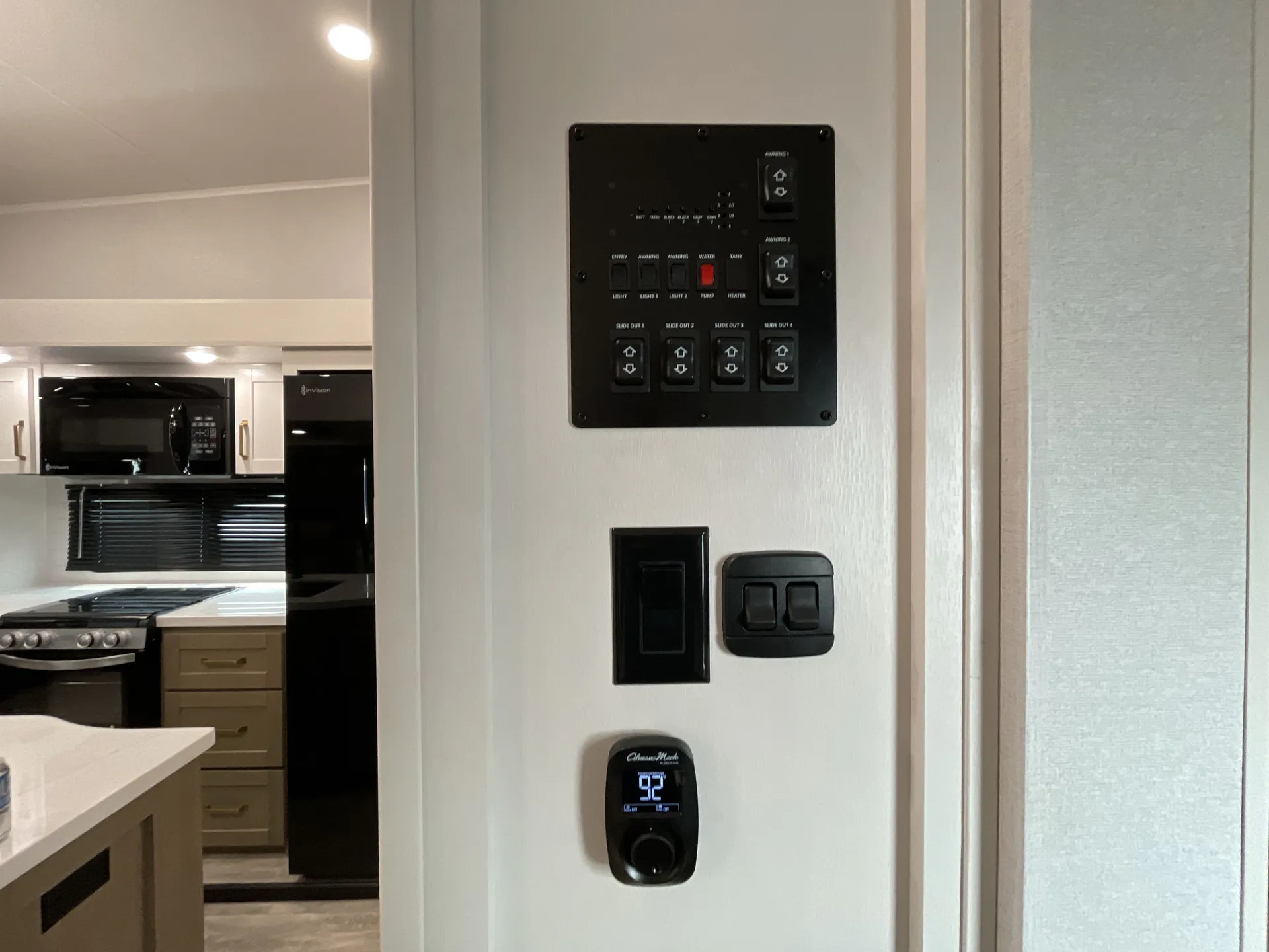 2027 Jayco Eagle 355MBQS — RV control panel and monitor beside a visible kitchen with refrigerator, microwave, range, and countertop