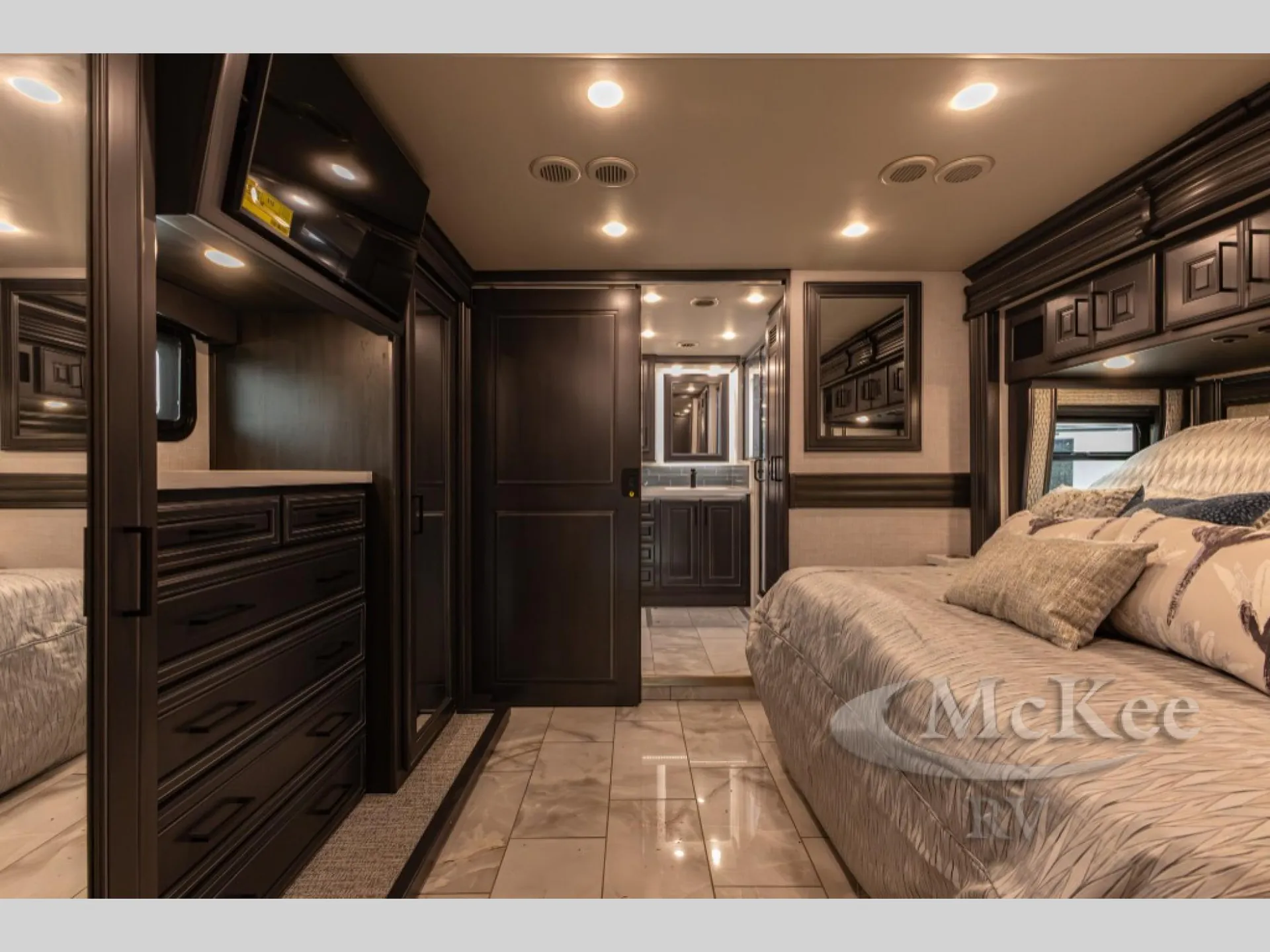 2027 Fleetwood Discovery LXE 40M — Main bedroom with a large bed, dresser, overhead cabinets, television, and bathroom visible through the doorway